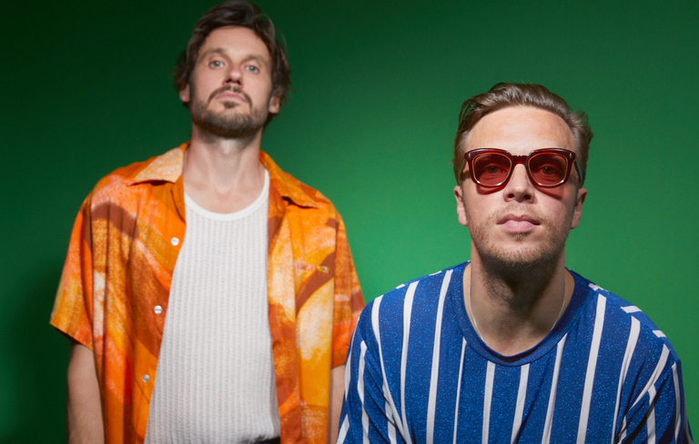 Promotional portrait of British indie band Friendly Fires featuring members posing against a vivid green backdrop in colorful shirts.